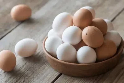A wooden bowl filled with white and brown eggs sits on a wooden table, with a few eggs scattered nearby.