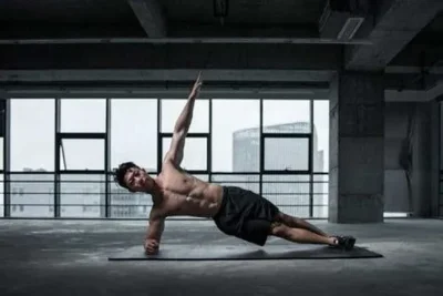 A shirtless man in black shorts does a side plank on a yoga mat in a spacious, industrial-style gym with large windows and city buildings visible outside.